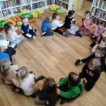 na zdjęciu widać dzieci siedzące na podłodze w sali bibliotecznej masujące sobie plecki.