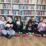 na zdjęciu widać dzieci siedzące na podłodze trzymające prace plastyczne chmurki ze słoneczkiem. dzieci siedzą na tle regałów z książkami.