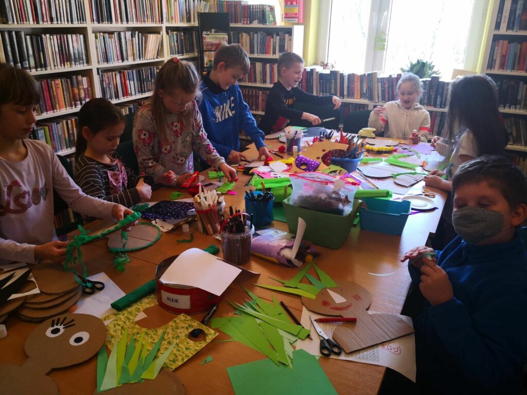 na zdjęciu widoczna jest sala biblioteczna, dzieci wokół stołu wykonują pracę plastyczną