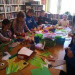 na zdjęciu widoczna jest sala biblioteczna, dzieci wokół stołu wykonują pracę plastyczną
