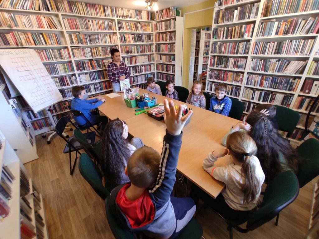 na zdjęciu widoczna jest sala biblioteczna, regały z książkami, na środku sali stoi stół, wokół stołu siedzą dzieci, bibliotekarz stoi przy stole w rękach trzyma książkę