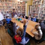 na zdjęciu widoczna jest sala biblioteczna, regały z książkami, na środku sali stoi stół, wokół stołu siedzą dzieci, bibliotekarz stoi przy stole w rękach trzyma książkę