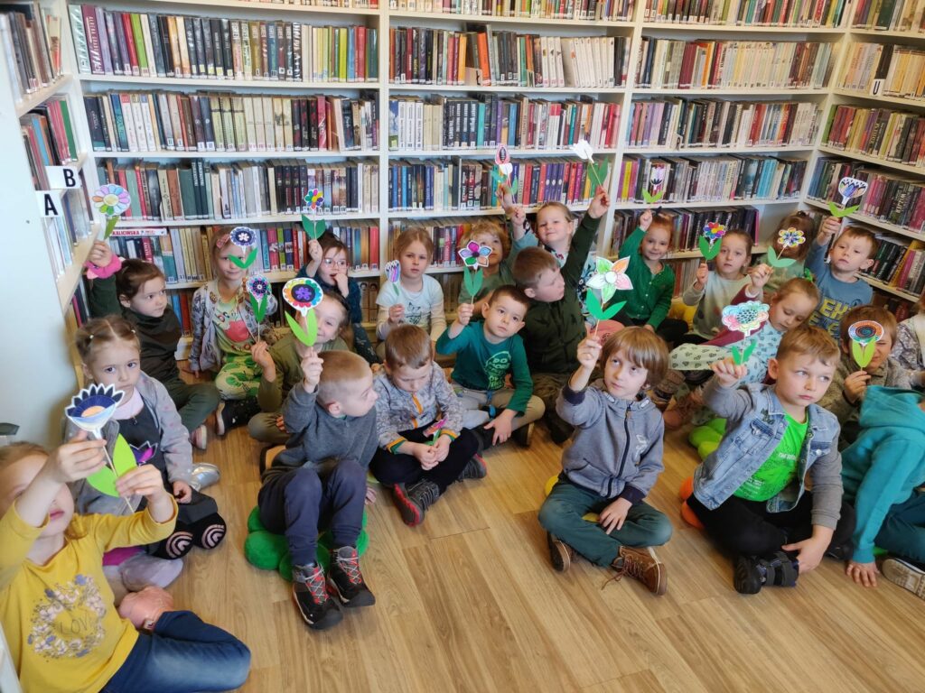 na zdjęciu widać salę biblioteczną, regały z książkami, na podłodze siedzą dzieci, w rękach trzymają kwiaty wykonane w czasie zajęć