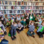 na zdjęciu widać salę biblioteczną, regały z książkami, na podłodze siedzą dzieci, w rękach trzymają kwiaty wykonane w czasie zajęć