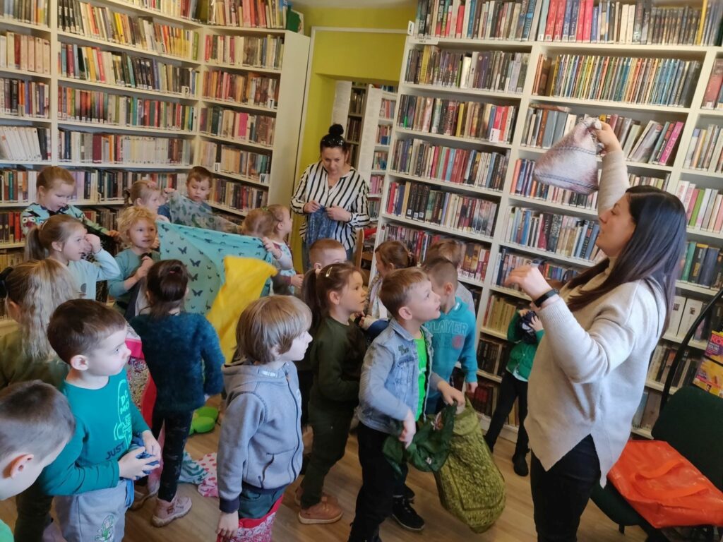 na zdjęciu widać salę biblioteczną, regały z książkami, na środku sali stoją dzieci i bibliotekarz, w dłoniach trzymają chusty