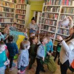 na zdjęciu widać salę biblioteczną, regały z książkami, na środku sali stoją dzieci i bibliotekarz, w dłoniach trzymają chusty