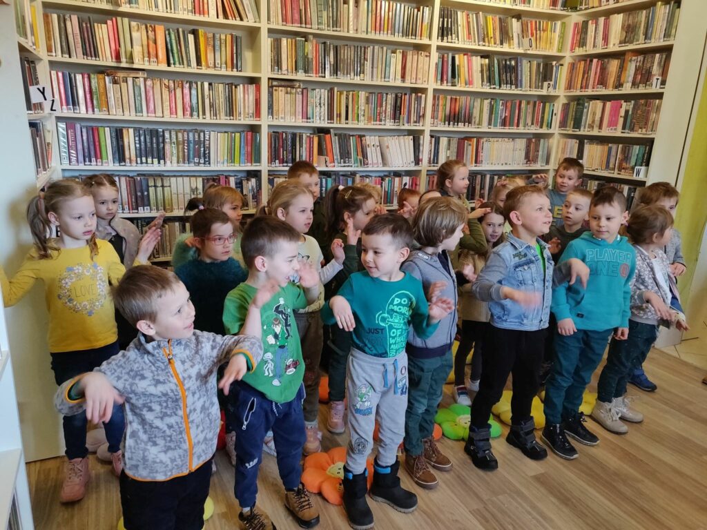 na zdjęciu widać salę biblioteczną, regały z książkami, na środku sali stoją przedszkolaki