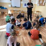 W szkolnej klasie dzieci siedzą na podłodze i wykonują pracę plastyczną i równocześnie słuchają czytanej im przez bibliotekarza bajki o niedźwiedziu.