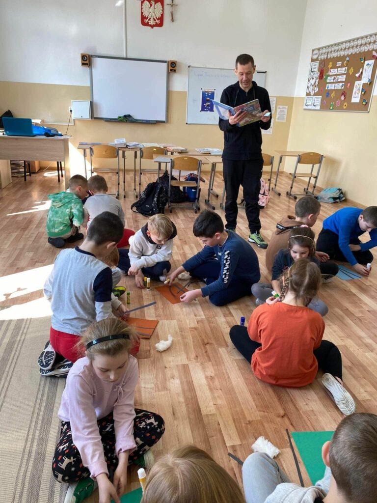 W szkolnej klasie dzieci siedzą na podłodze i wykonują pracę plastyczną i równocześnie słuchają czytanej im przez bibliotekarza bajki o niedźwiedziu.