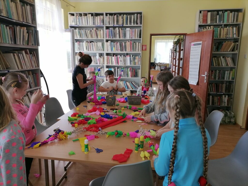 Na zdjęciu widoczne są dzieci (3 chłopców i 6 dziewczynek) uczestniczące w wielkanocnych warsztatach w bibliotece. Sześcioro dzieci stoi, a troje kuca na tle regałów z książkami i prezentują palmy wielkanocne wykonane podczas warsztatów. Palmy wykonane są z bibuły i krepiny.