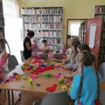 Na zdjęciu widoczne są dzieci (3 chłopców i 6 dziewczynek) uczestniczące w wielkanocnych warsztatach w bibliotece. Sześcioro dzieci stoi, a troje kuca na tle regałów z książkami i prezentują palmy wielkanocne wykonane podczas warsztatów. Palmy wykonane są z bibuły i krepiny.