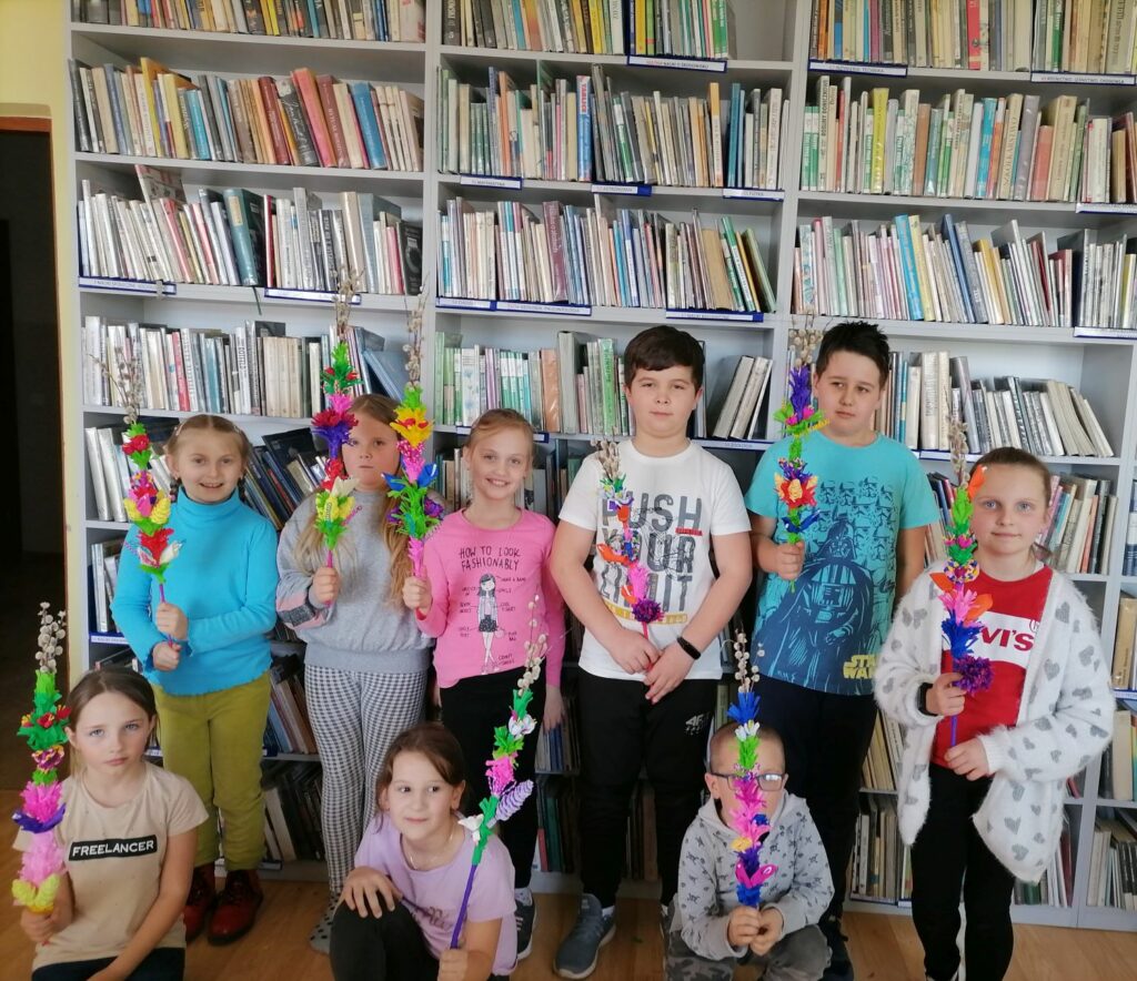 Na zdjęciu widoczne są dzieci wykonujące palmy wielkanocne podczas warsztatów odbywających się w bibliotece. W tle regały z książkami.
