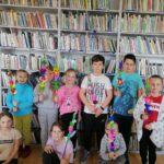 Na zdjęciu widoczne są dzieci wykonujące palmy wielkanocne podczas warsztatów odbywających się w bibliotece. W tle regały z książkami.