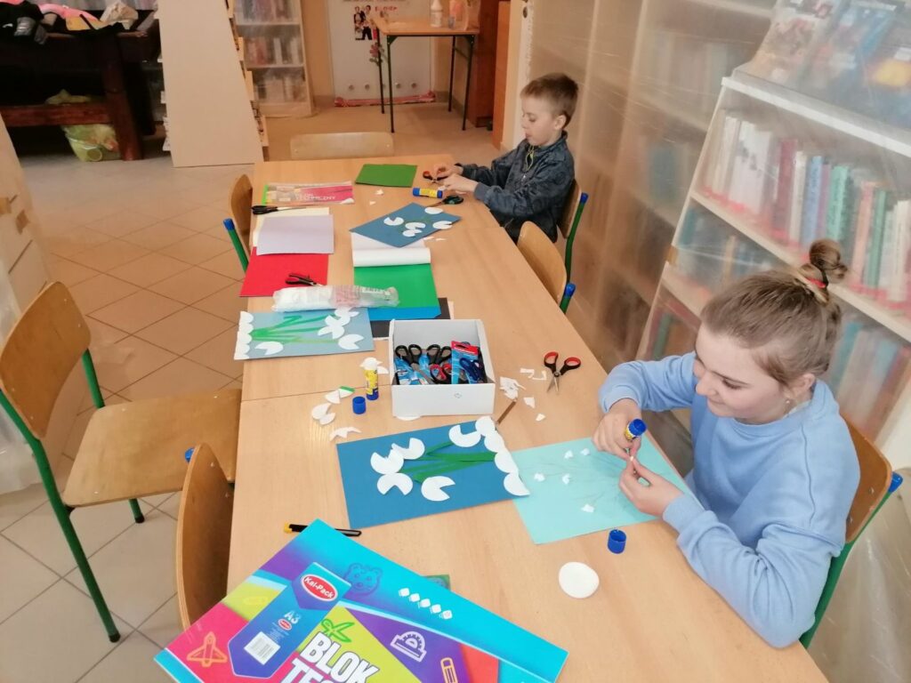 Dzieci siedzą przy bibliotecznym stole i przy pomocy kolorowego papieru, kleju, nożyczek i płatków kosmetycznych wykonują na kartkach przebiśniegi.