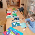 Dzieci siedzą przy bibliotecznym stole i przy pomocy kolorowego papieru, kleju, nożyczek i płatków kosmetycznych wykonują na kartkach przebiśniegi.