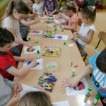 Na zdjęciu widoczna jest grupa przedszkolaków (4 chłopców, 7 dziewczynek) wykonujących przy stolikach pracę plastyczną przedstawiającą kolorowe muffinki z plasteliny, falistej tekturki oraz posypek.
