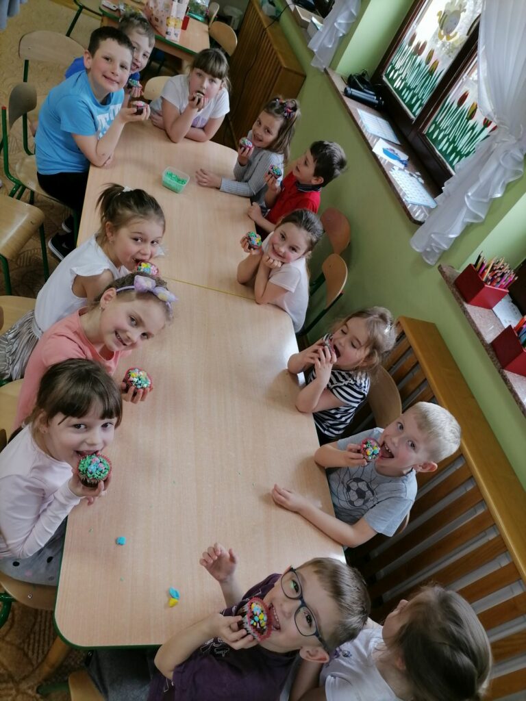 Na zdjęciu widoczna jest grupa przedszkolaków siedzących przy stoliku i jedzących ozdobione przez siebie muffinki.