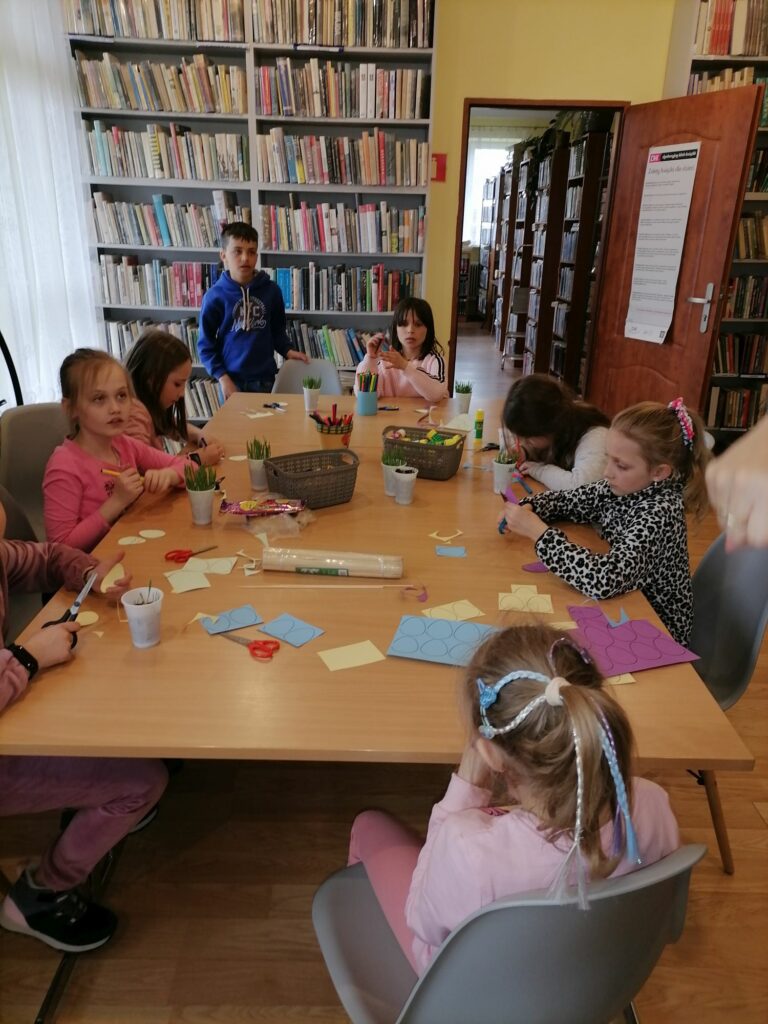 Na zdjęciu widoczna jest grupa dzieci uczestniczących w warsztatach wielkanocnych. Dzieci siedzą przy stolikach i wykonują pisankowe pikery, które później umieszczą w kubeczkach z posianym owsem. W tle regały z książkami.