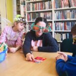 na zdjęciu widać stół i siedzące przy nim dzieci i bibliotekarkę, która ma na nosie ogromne okulary.
