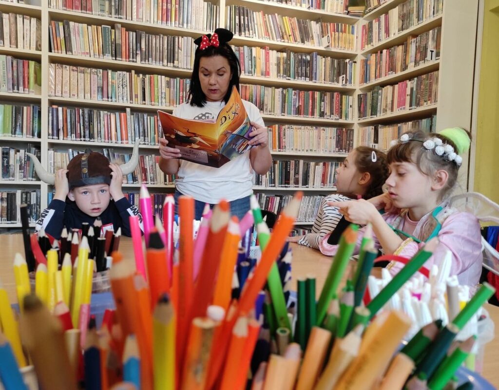 na zdjęciu widać bibliotekarkę czytającą dzieciom, na pierwszym planie widać kolorowe kredki.