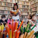 na zdjęciu widać bibliotekarkę czytającą dzieciom, na pierwszym planie widać kolorowe kredki.
