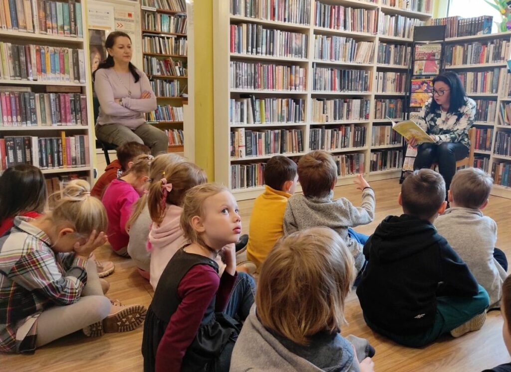 na zdjęciu widać sale biblioteczną, regały z książkami i dzieci siedzące na podłodze i słuchające czytającej bibliotekarki.