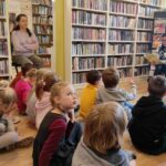 na zdjęciu widać sale biblioteczną, regały z książkami i dzieci siedzące na podłodze i słuchające czytającej bibliotekarki.