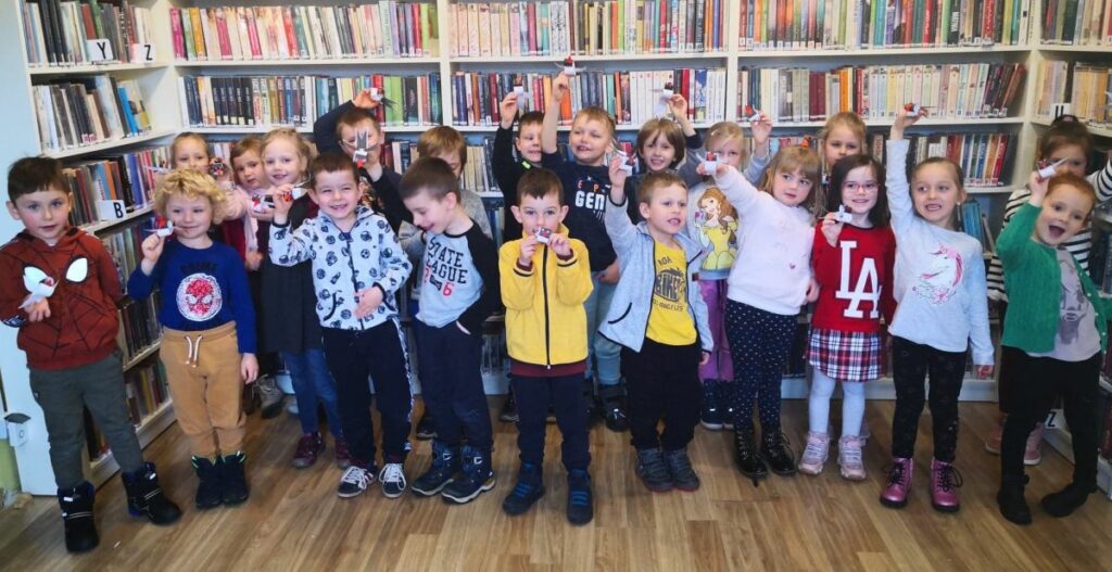 na zdjęciu widać dzieci stojące na tle regałów z książkami i trzymające wykonane przez siebie papierowe jaskólki.