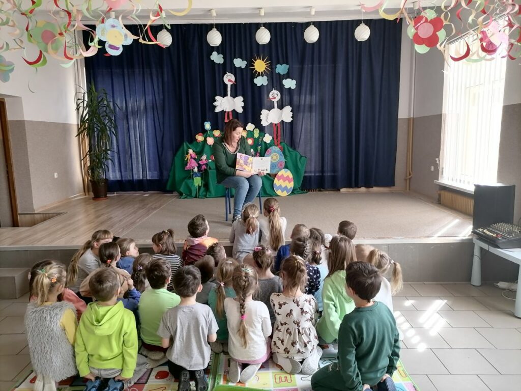 Na zdjęciu widoczna jest grupa przedszkolaków siedzących na matach i słuchających bajki czytanej im przez bibliotekarza. W tle dekoracje wiosenne: bociany, kwiaty, pisanki itp.