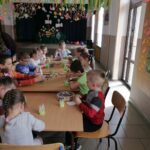 Na zdjęciu widoczna jest grupa przedszkolaków siedzących przy stolikach i wykonujących prace plastyczne. W tle dekoracje wiosenne.