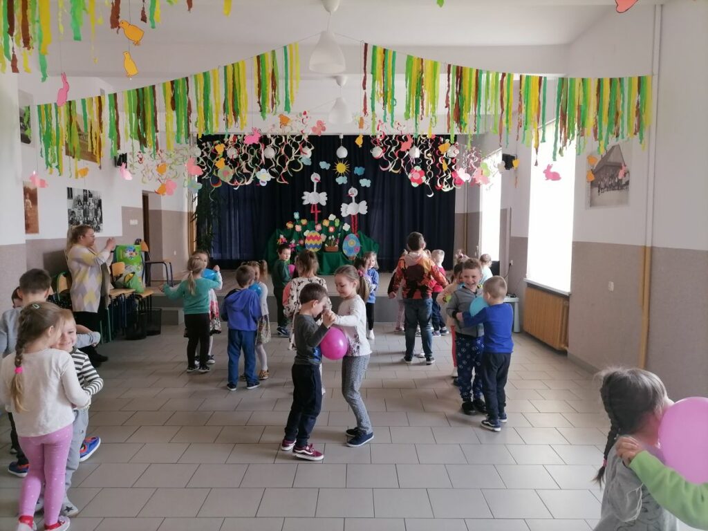 Na zdjęciu widoczna jest grupa przedszkolaków uczestniczących w zabawach ruchowych - "Zajączkowym tańcu z balonikiem" polegającej na tańcu w parach z umieszczonym między osobami balonikiem. Zabawę wygrywa ta osoba, której balonik nie spadnie na ziemię.
