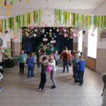 Na zdjęciu widoczna jest grupa przedszkolaków uczestniczących w zabawach ruchowych - "Zajączkowym tańcu z balonikiem" polegającej na tańcu w parach z umieszczonym między osobami balonikiem. Zabawę wygrywa ta osoba, której balonik nie spadnie na ziemię.