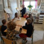 W sali bibliotecznej grupa dzieci siedzi przy stole i słucha z uwagą czytanej im przez pana bibliotekarza książki o zagadkach i tajemnicach.