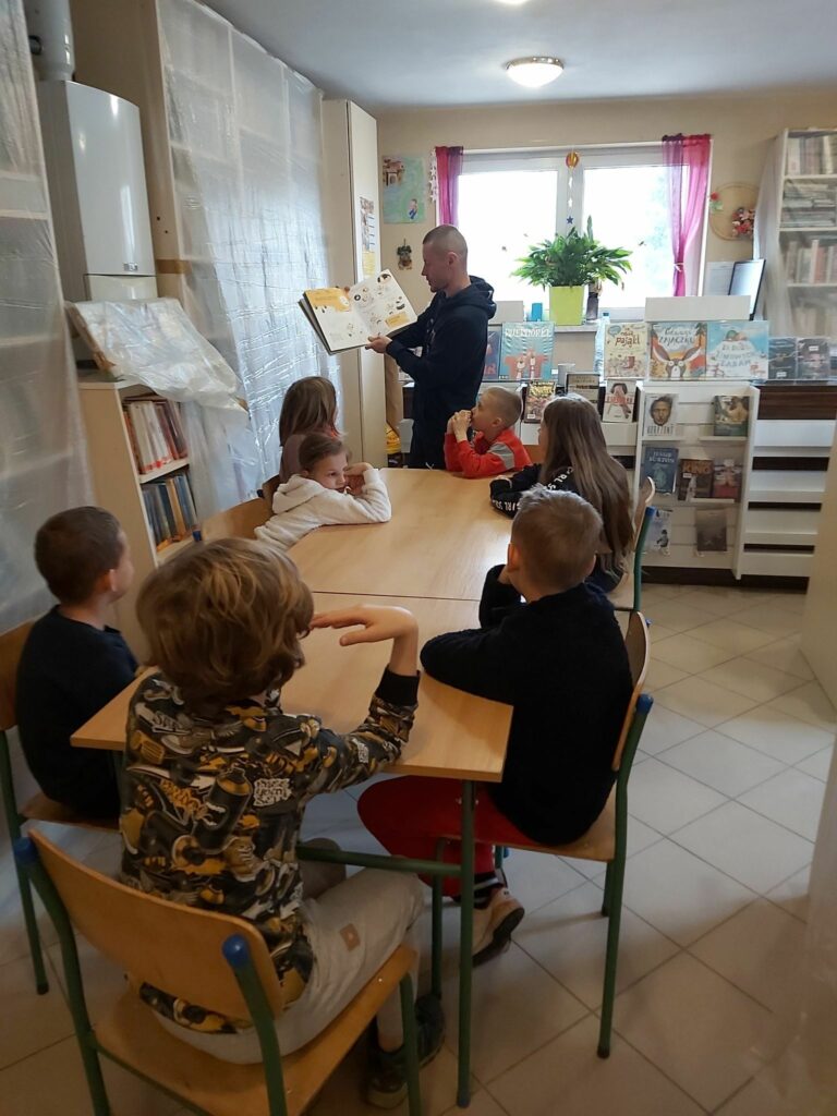 W sali bibliotecznej grupa dzieci siedzi przy stole i słucha z uwagą czytanej im przez pana bibliotekarza książki o zagadkach i tajemnicach.