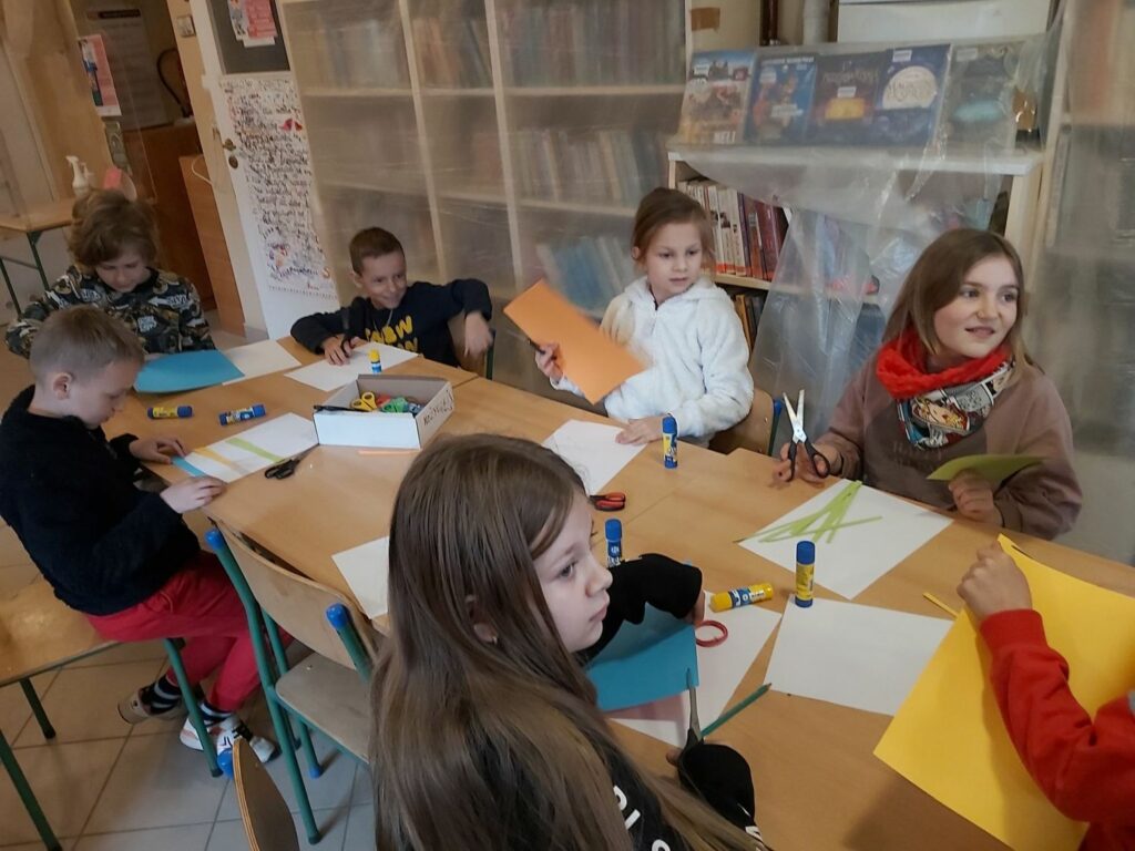 Mali uczestnicy spotkania siedzą przy stole i wycinają z papieru kolorowe paski, którymi później udekorują swoje kartki świąteczne.