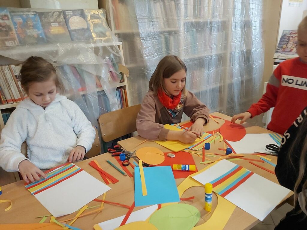 Dwie siedzące przy stole dziewczynki pracują w skupieniu. Jedna z nich przykleja na kartce kolorowe paski papieru, druga natomiast rysuje wielkie wielkanocne jajo.