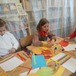 Dwie siedzące przy stole dziewczynki pracują w skupieniu. Jedna z nich przykleja na kartce kolorowe paski papieru, druga natomiast rysuje wielkie wielkanocne jajo.