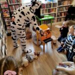 na zdjęciu widać dzieci siedzące na podłodze w bibliotece oraz bibliotekarkę przebrana w biło czarny strój krowy
