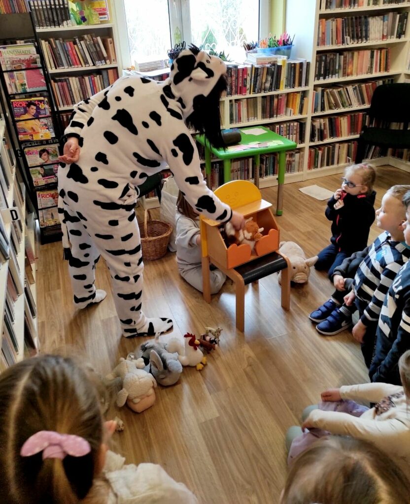 na zdjęciu widać dzieci siedzące na podłodze w bibliotece oraz bibliotekarkę przebrana w biło czarny strój krowy