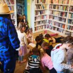 na zdjęciu widać sale biblioteczną, regały z książkami, dzieci na podłodze kolorujące obrazek oraz stojącą bibliotekarkę przebraną za farmera.