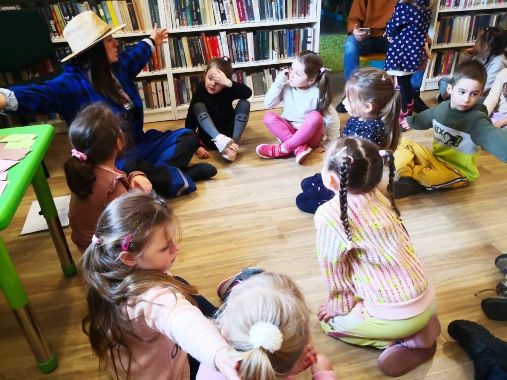 na zdjęciu widać dzieci siedzące na podłodze a wraz nimi siedzi bibliotekarka przebrana za rolnika.