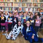 na zdjęciu widać salę biblioteczną. Na tle regałów stoją dzieci trzymające pomalowane przez siebie kolorowe kurki. Przed dziećmi siedzą bibliotekarki . Jedna w przebraniu biało czarnej krowy, druga w przebraniu rolnika w słomkowym kapeluszu.