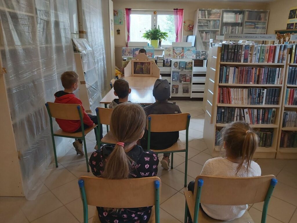 W bibliotecznej sali dzieci siedzą na krzesłach i oglądają prezentowany im przez bibliotekarza teatrzyk obrazkowy Kamishibai.