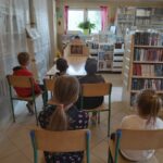 W bibliotecznej sali dzieci siedzą na krzesłach i oglądają prezentowany im przez bibliotekarza teatrzyk obrazkowy Kamishibai.