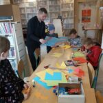 Przy wielkim stole grupa dzieci wycina z kolorowego papieru różne postacie, w tym czasie bibliotekarz czyta im bajkę pt. „Wiewiórki, które nie chciały się dzielić”.