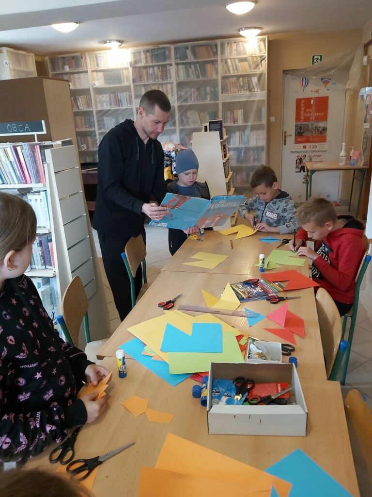 Przy wielkim stole grupa dzieci wycina z kolorowego papieru różne postacie, w tym czasie bibliotekarz czyta im bajkę pt. „Wiewiórki, które nie chciały się dzielić”.