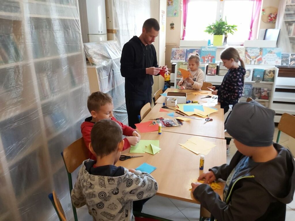 Grupa dzieci razem z panem bibliotekarzem pracują przy stole nad swoimi zakładkami do książek. Do ich zdobienia mali artyści używają kolorowego papieru, flamastrów i własnej wyobraźni.