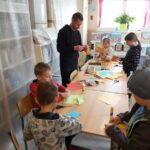 Grupa dzieci razem z panem bibliotekarzem pracują przy stole nad swoimi zakładkami do książek. Do ich zdobienia mali artyści używają kolorowego papieru, flamastrów i własnej wyobraźni.