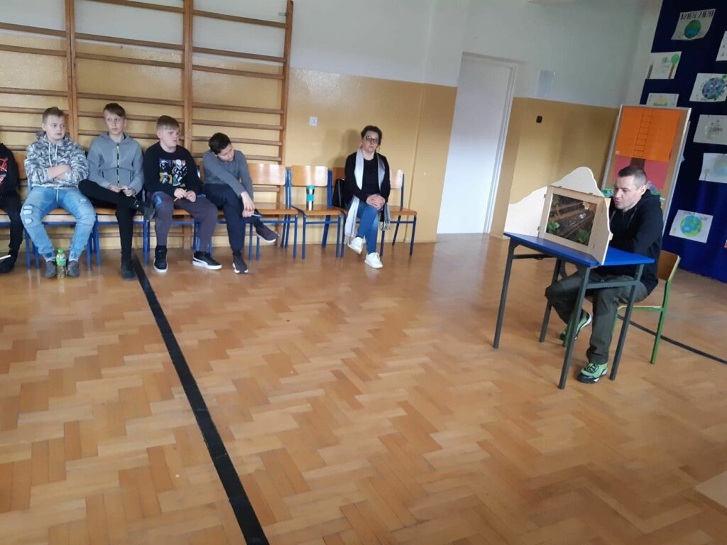W szkolnej sali gimnastycznej trwa przedstawienie. Zgromadzeni uczniowie oglądają teatrzyk obrazkowy Kamishibai pt. „Jak żyje Ziemia”, który demonstruje im bibliotekarz.