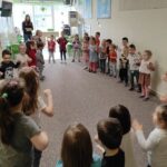 na zdjęciu widać salę przedszkolną w której w kole stoją ułożone dzieci a wśród nich stoją bibliotekarka i przedszkolanka.